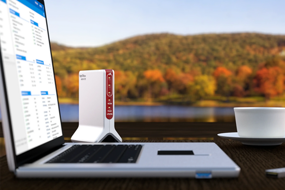 Laptop mit Router am Ufer eines Sees, umgeben von herbstlichen Bäumen und einer Tasse Kaffee.