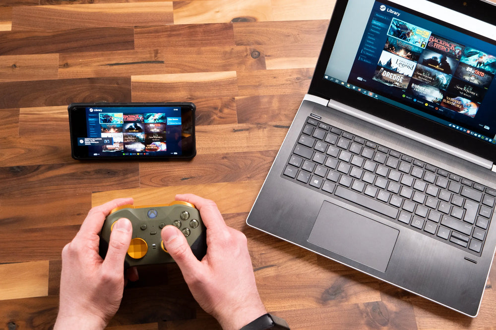 Person spielt Videospiele mit Gamecontroller, Laptop und Smartphone auf einem Holztisch.