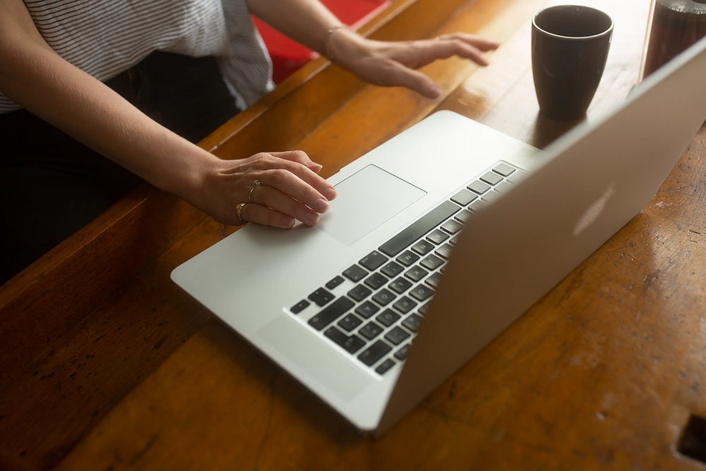 Person arbeitet an einem Laptop auf einem Holztisch, mit einer Tasse Kaffee daneben.