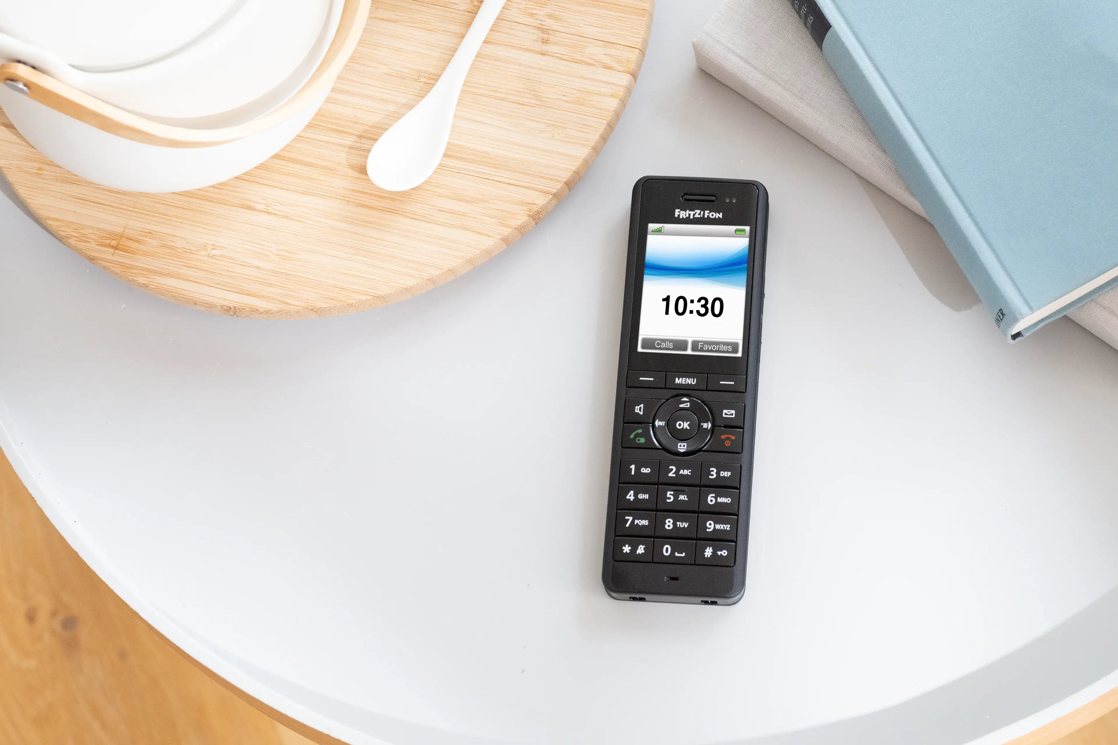 Schwarzes Festnetztelefon auf einem Tisch, Uhrzeit 10:30, umgeben von Geschirr und einem Buch.