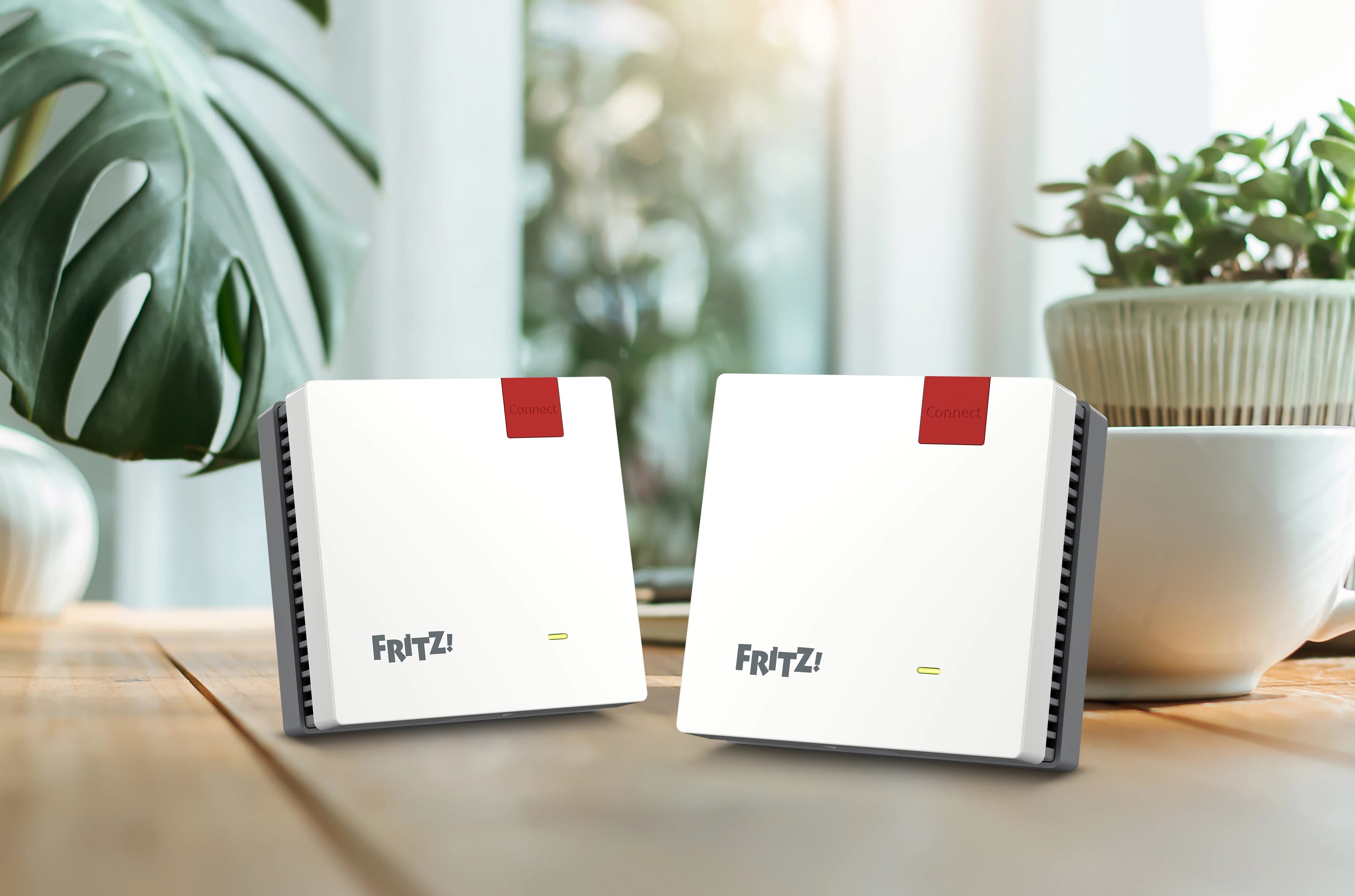 Zwei FRITZ!Box-Router auf einem Holztisch, umgeben von Pflanzen, mit natürlichem Licht.