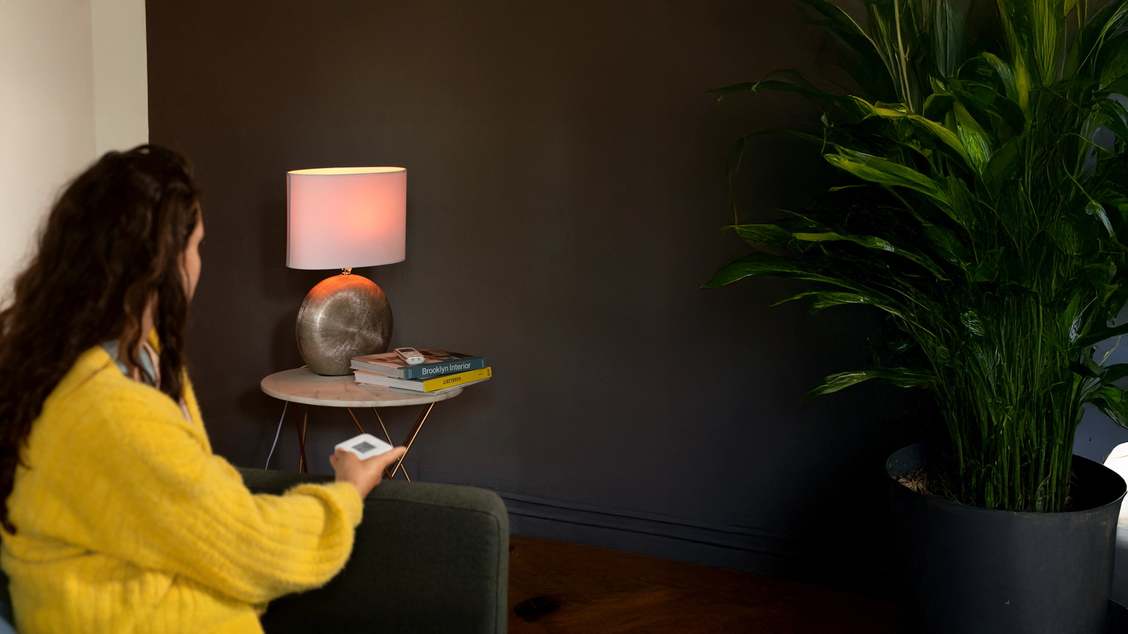 Frau sitzt auf der Couch in gelbem Pullover, greift zur Fernbedienung neben einer Lampe und einer Pflanze.