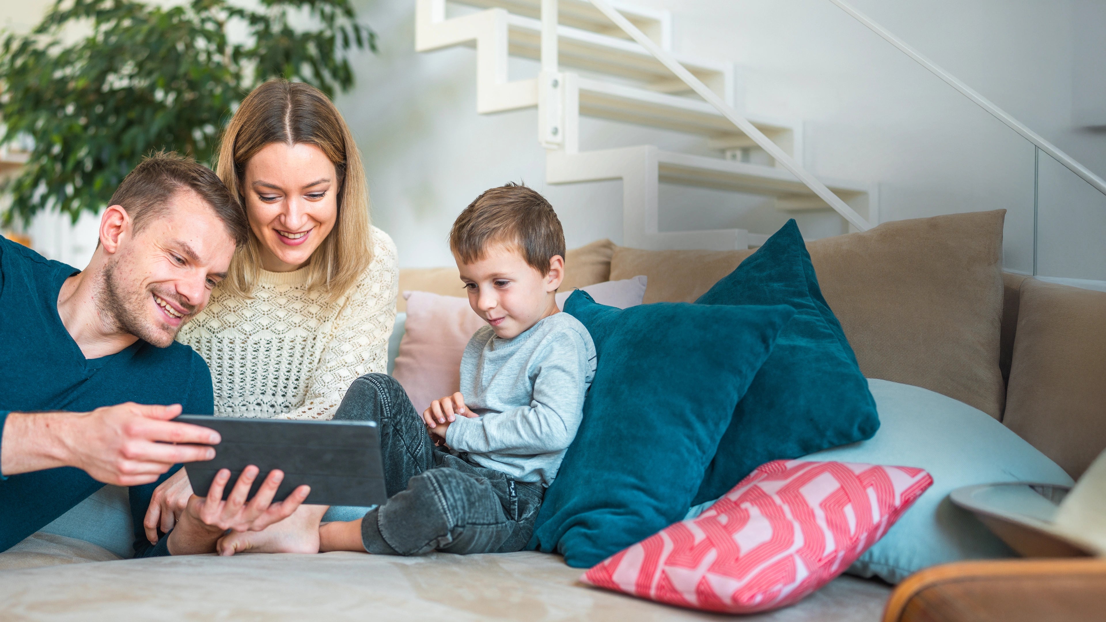 Familie betrachtet gemeinsam ein Tablet auf der Couch mit bunten Kissen in einem modernen Wohnraum.